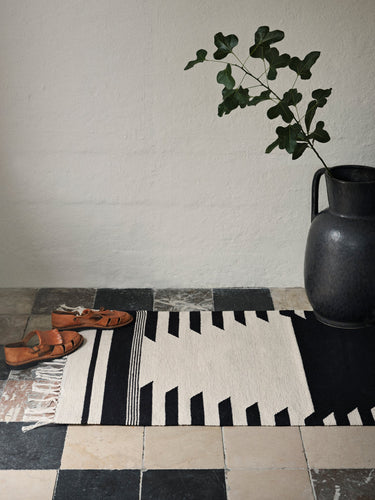 Alfombra de algodón blanco roto y negro de Broste Copenhagen junto a un jarrón negro