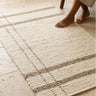 Alfombra Duna cruda a rayas topo sobre suelo claro de piedra, junto a una silla de madera y unos pies descalzos