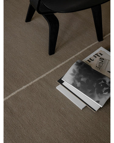 Alfombra de lana y lino verde con línea central de Linie Design: detalle de la línea junto a una silla y un libro.