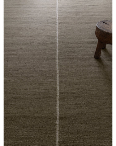 Alfombra de lana y lino verde con línea central de Linie Design: vista del tejido junto a un taburete.