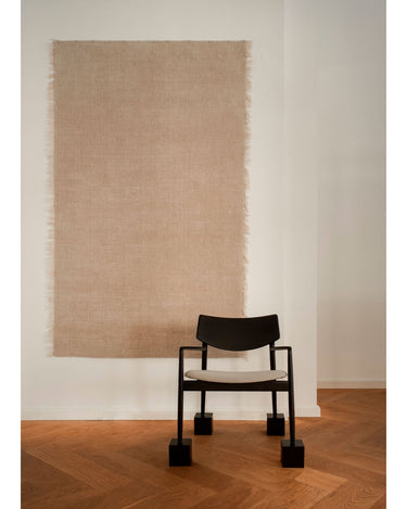 Alfombra de lana marfil con flecos de lino de Linie Design: colgada en una pared junto a una silla.