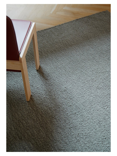 Alfombra de lana texturada Pebbled Poem de Linie Design, en tono verde grisáceo, junto a una silla
