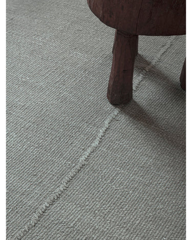 Alfombra de lino gris menta con líneas orgánicas de Linie Design: detalle del tejido bajo las patas del taburete.