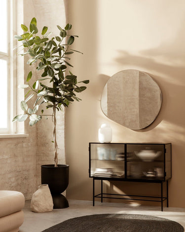 Aparador de acero negro con cristal estriado de Ferm Living: aparador de dos puertas de cristal bajo espejo redondo, con planta a un lado y objetos dentro.
