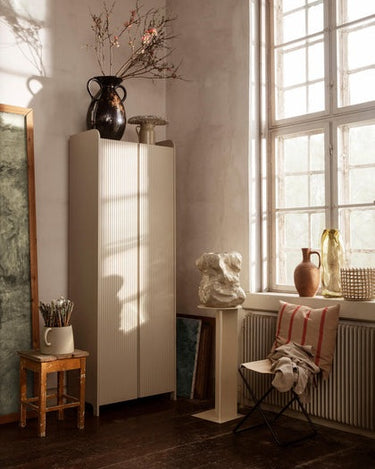 Armario alto de puertas acanaladas de Ferm Living, MDF en beige claro: armario beige junto a una ventana, un taburete y un pedestal.