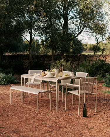 Banco de Ferm Living: banco beige junto a mesa y sillas de jardín, sobre grava roja y bajo árboles.