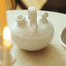 Botijo de gres blanco con decoración en relieve de Leandro Cano: botijo blanco con asa y dos bocas sobre mesa de tejido natural.