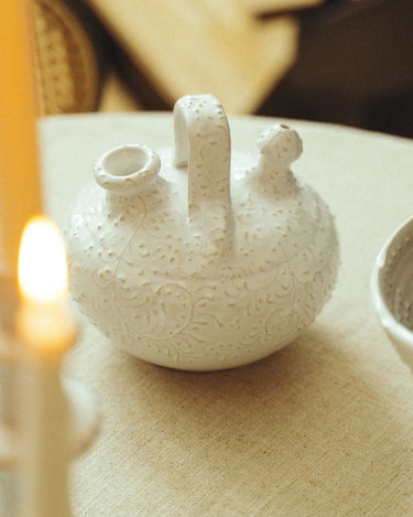 Botijo de gres blanco con decoración en relieve de Leandro Cano: botijo blanco con asa y dos bocas sobre mesa de tejido natural.