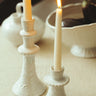 Candelabro alto de gres blanco con relieves de Leandro Cano: candelabro alto blanco con vela, junto a otro bajo sobre mesa.