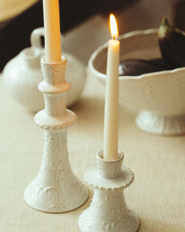 Candelabro alto de gres blanco con relieves de Leandro Cano: candelabro alto blanco con vela, junto a otro bajo sobre mesa.