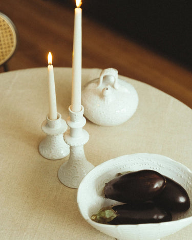 Candelabro bajo de gres blanco con relieves de Leandro Cano: candelabro bajo y alto con velas encendidas, botijo y frutero sobre mesa.