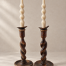 Candeleros antiguos de madera torneada oscura en pareja, colocados uno junto al otro con velas en espiral color crema sobre fondo topo claro