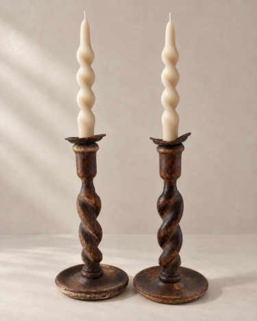 Candeleros antiguos de madera torneada oscura en pareja, colocados uno junto al otro con velas en espiral color crema sobre fondo topo claro