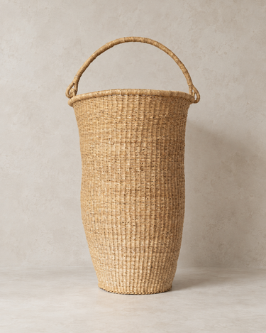 Cesta de fibras naturales de Matuta Home con el asa levantada sobre fondo de cemento topo claro
