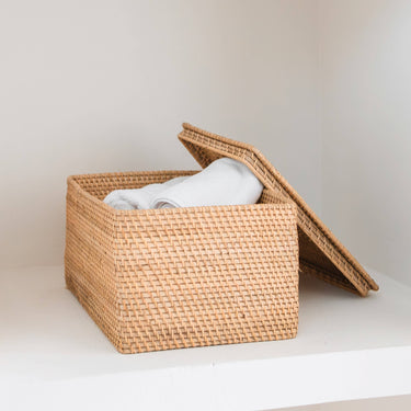 Cesta rectangular de fibras naturales de Soeji con tapa entreabierta y textiles blancos en el interior