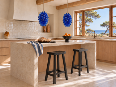 Cocina mediterránea con isla de piedra, dos lámparas Uva azules de vidrio, taburetes negros y naranjas de cerámica de Matuta Home.