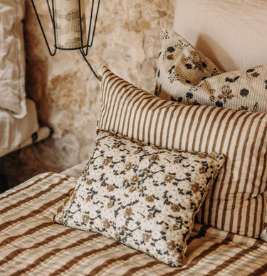 Cojín de algodón con estampado block print en verde oliva y crudo de Le Monde Sauvage, junto a cojines de rayas sobre una cama