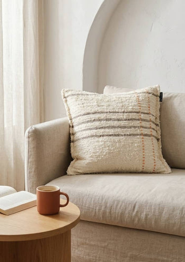 Cojín Aura a rayas sobre un sofá crudo junto a una mesa de madera y una ventana