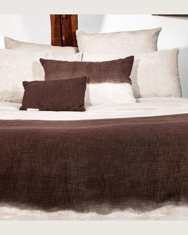 Cojín marrón de algodón degradado de Bed and Philosophy, sobre una cama con otros cojines y un plaid