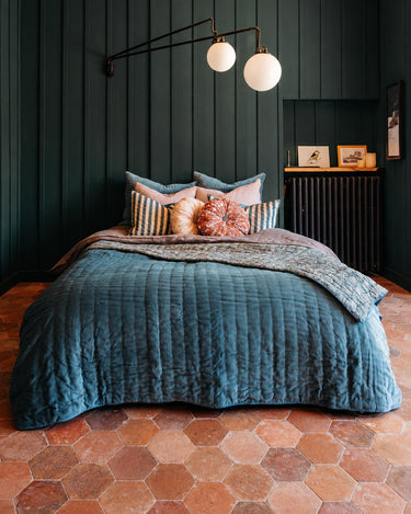 Cojín rosa floral de Le Monde Sauvage en una cama con textiles azules y cojines de rayas