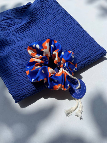 Coletero de House of Scrunchies: coletero sobre un tejido azul de punto, modelo azul y naranja