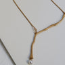 Collar dorado sinuoso con perla de vidrio de Joidart: collar dorado de cadena y motivo sinuoso en forma de Y con extremo blanco nacarado, sobre plano gris.