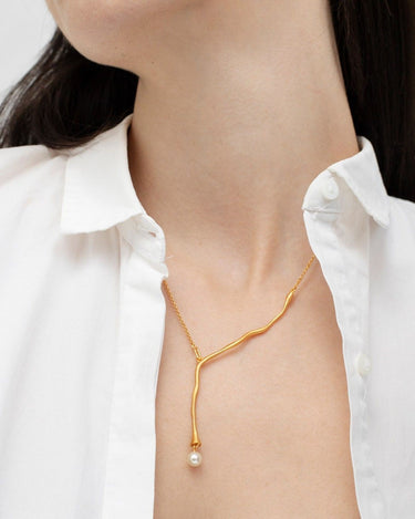 Uno de los collares dorados sinuosos Eclat de Joidart, con extremo blanco y llevado con camisa blanca abierta.