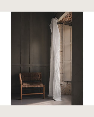 Cortina de lino blanco con pespunte vertical, colgada en un interior oscuro junto a una silla