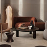 Diván izquierdo de tejido de lana marrón rojizo de Ferm Living: ambiente interior: diván marrón rojizo bajo luz lateral, mesa redonda oscura delante, lámpara alta clara y varios asientos alrededor.