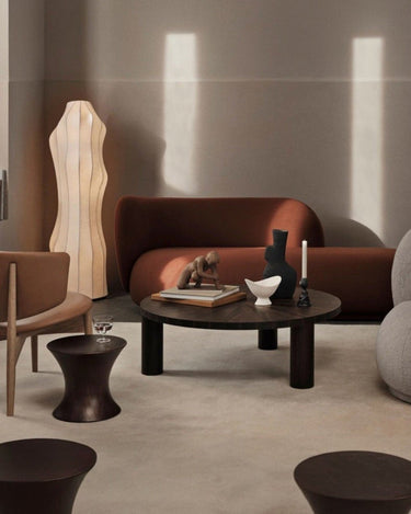Diván izquierdo de tejido de lana marrón rojizo de Ferm Living: ambiente interior: diván marrón rojizo bajo luz lateral, mesa redonda oscura delante, lámpara alta clara y varios asientos alrededor.