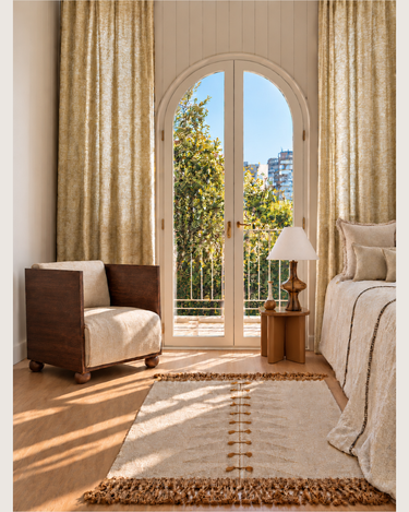 Dormitorio con textiles claros, cortinas y una butaca junto a una ventana en arco
