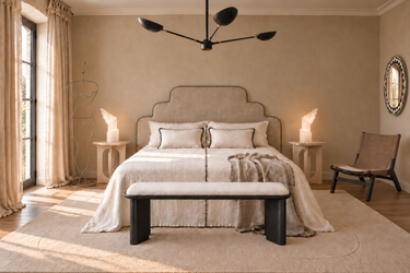 Dormitorio Matuta Home con cabecero de lino, mesitas de travertino, banco negro, sillón de cuero y espejo geométrico. Imagen generada con IA.