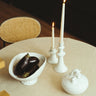 Frutero ovalado de gres blanco con pie de Leandro Cano: frutero con berenjenas junto a botijo y dos candelabros con velas encendidas.