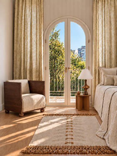 Dormitorio en tonos naturales con butaca de madera oscura, mesa auxiliar caramelo, lámpara de vidrio y soliflor cerámico junto a una ventana en arco.