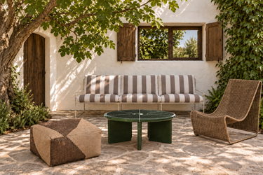 Patio bajo una higuera con sofá Desert a rayas, mesa Deya verde oscuro, sillón Calantha de ratán y puf Forene.