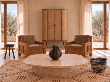 Salón mediterráneo con sillones de madera, armario de mango y mimbre, mesa de travertino, cortinas claras, taburete oscuro, jarrón negro y cesta Bolga.