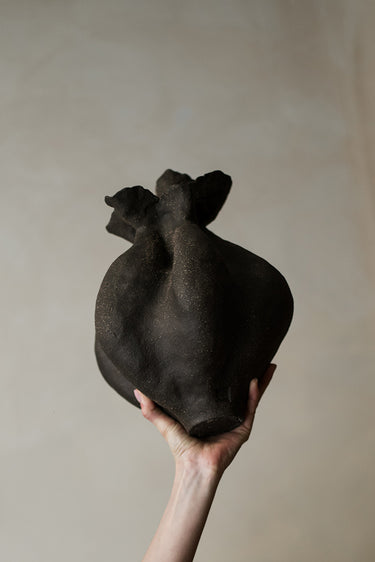 Jarrón negro de cerámica artesanal con cuerpo lobulado y boca irregular, sostenido en la mano.