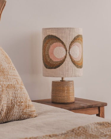 Lámpara de mesa con pie cerámico marrón y pantalla multicolor de âme philo, junto a una cama sobre una mesa de madera