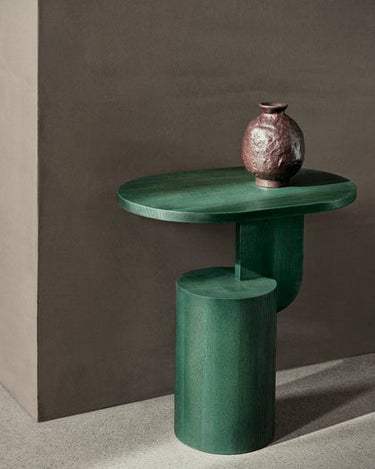 Mesa auxiliar de fresno con sobre ovalado de Ferm Living, fresno verde oscuro: mesa auxiliar verde con jarrón cerámico junto a pared.