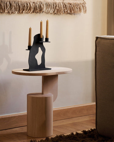 Mesa auxiliar ovalada de fresno macizo natural de Ferm Living junto a un sillón, con candelabro negro.