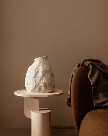 Mesa auxiliar de fresno con jarrón blanco junto a un sillón marrón.