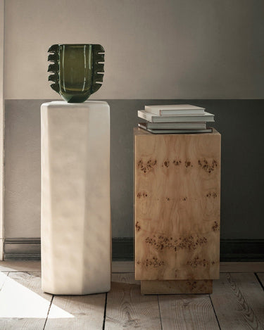 Mesa de Ferm Living: pedestal con libros junto a otra peana clara y objeto verde, ante pared con molduras.