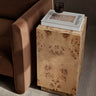 Mesa auxiliar de raíz de álamo con almacenaje de Ferm Living: pedestal de madera veteada junto a asiento tapizado marrón parcialmente visible, con libros y vaso encima.