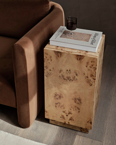 Mesa auxiliar de raíz de álamo con almacenaje de Ferm Living: pedestal de madera veteada junto a asiento tapizado marrón parcialmente visible, con libros y vaso encima.