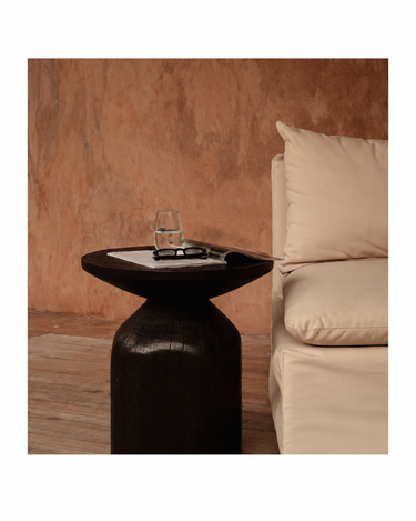 Mesa auxiliar redonda de madera oscura, con un vaso y un libro junto a un asiento