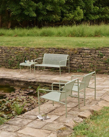 Mesa de Ferm Living: mesa baja junto a sofá y sillones verdes en jardín con estanque y muro de piedra.