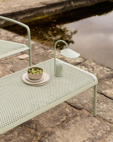 Mesa de centro de jardín rectangular de acero perforado de Ferm Living: mesa baja rectangular verde junto al agua, con platos, uvas y lámpara portátil encima.