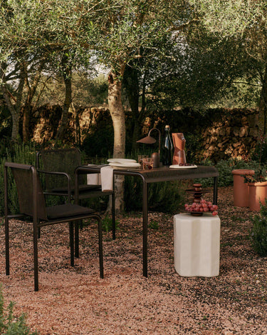 Mesa de Ferm Living: mesa de comedor marrón con sillas en jardín arbolado, sobre suelo de grava.