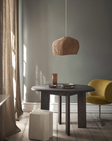 Mesa de haya oscura bajo una lámpara de fibras, con silla amarilla y taburete blanco.