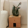 Mesita de noche de chapa de roble con un cajón de Ferm Living: mesita de un cajón junto a cama clara, con jarrón verde ondulado y ramas.